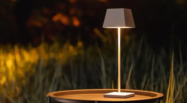 Nowoczesna lampa na balkon, stojąca na stoliku, oświetla wieczorny ogród.