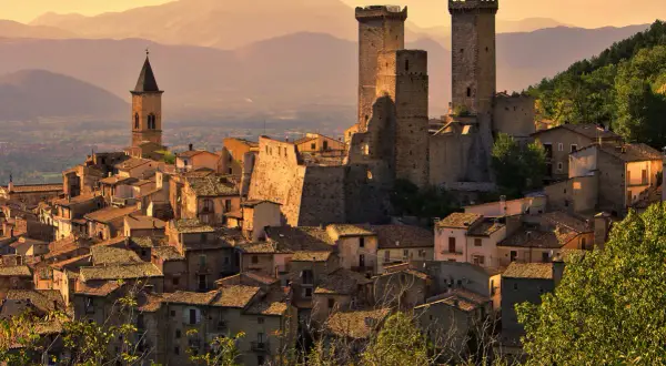 Borghi da visitare in Abruzzo: un castello medievale domina un villaggio arroccato, con torri imponenti e case in pietra sotto un cielo dorato.