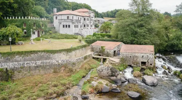 Antigua barcala con molinos de piedra junto a un río caudaloso, con una casa señorial cubierta de hiedra al fondo.