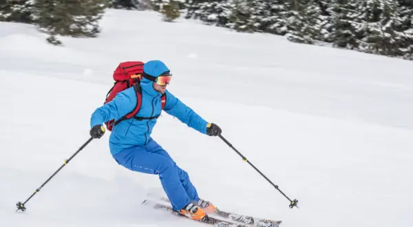 Narciarz w niebieskiej kurtce i goglach zjeżdża po ośnieżonym stoku, niosąc czerwony plecak. To prawdziwy miłośnik ski tury!