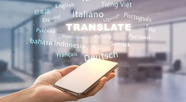 Dłoń trzyma smartfon, nad którym unosi się napis "TRANSLATE" otoczony nazwami języków. To pokazuje, jak przetłumaczyć tekst na wiele sposobów.