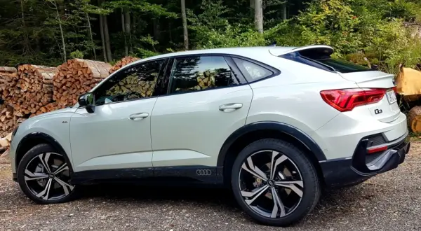 Nowy Audi Q3 2025 w kolorze białym, zaparkowany na leśnej drodze, z widocznymi stosami drewna w tle.