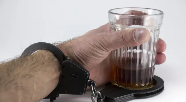 Ręka w kajdankach trzyma szklankę z alkoholem. To symbol zniewolenia, które może prowadzić do tego, dlaczego alkoholik chudnie.