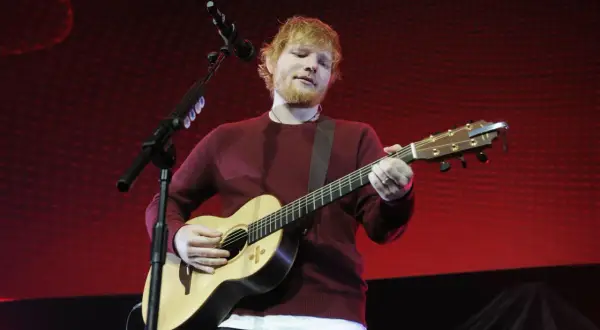 Ed Sheeran gra na gitarze akustycznej na tle czerwonej sceny.
