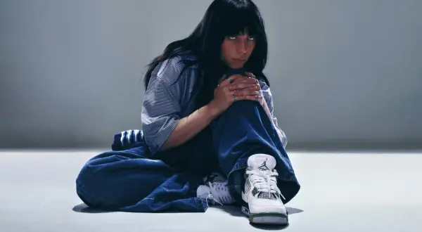 Billie Eilish w luźnych dżinsach i koszuli, zamyślona, w butach Jordan. Tekst "What was I made for?" pasuje do jej nastroju.