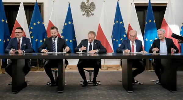 Pięciu mężczyzn w garniturach siedzi przy stole, podpisując dokumenty. W tle flagi Polski i UE. Może to być ważny moment dla sejmowej komisji śledczej.