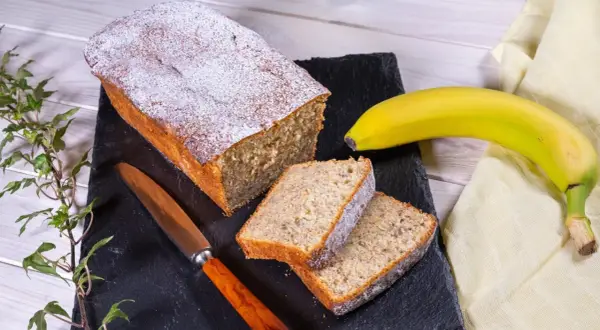 Ein saftiger Bananenkuchen, bestäubt mit Puderzucker, liegt auf einem schwarzen Brett. Daneben zwei Scheiben und eine reife Banane. Das ist der beste Bananenkuchen der Welt!