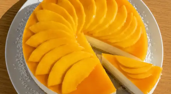 En krämig mango cheesecake med färska mangoskivor som pryder toppen. En bit är uppskuren och visar den vita fyllningen.