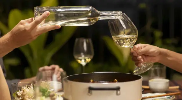Hälla upp vitt vin till en måltid med tomatsås. En flaska hälls i ett glas, med en gryta och mat i bakgrunden.