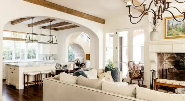A cozy living room with a fireplace and kitchen, featuring exposed wooden beams and arched doorways, embodying a warm mediterranean interior design.