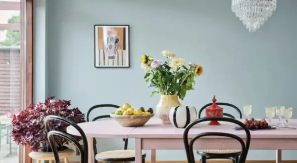 En matplats med ljusrosa bord, blommor i en gul vas och en skål med frukt. Kanske någon har valt att måla glasvasen för att få den att passa in.