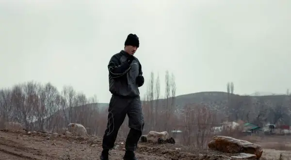 A boxer in a black tracksuit and beanie is running outdoors, practicing his footwork. The barren landscape suggests a cold day for this intense training.