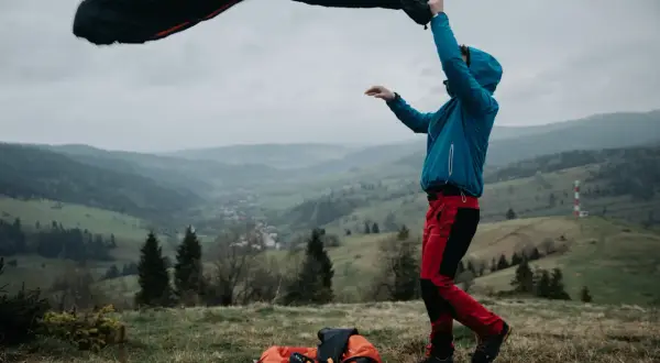 Osoba w niebieskiej kurtce i czerwonych spodniach rozkłada śpiwór puchowy czy syntetyczny na tle górskiego krajobrazu.