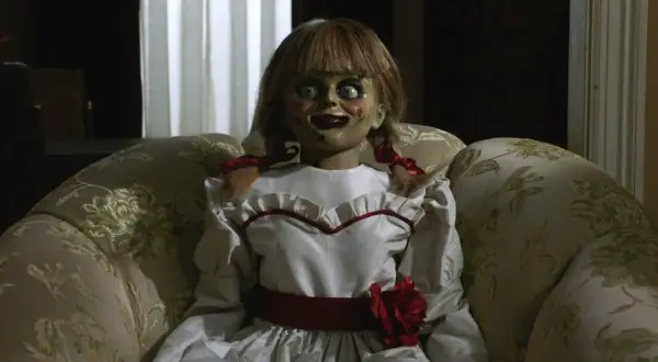 Den läskiga riktiga Annabelle dockan sitter i en blommig soffa, med sina stora ögon och röda rosett.