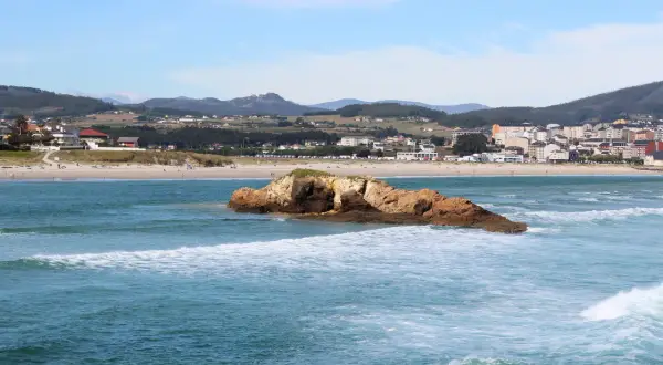 Olas rompen en la playa de Barreiros, con una roca en primer plano y el pueblo al fondo.