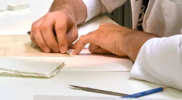 Main tenant avec précision un scalpel pour enlever de la colle sur un document ancien. Des outils et du papier sont disposés sur la table.