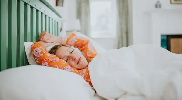 A woman in an orange patterned shirt stretches in bed, cozy under white sheets. She looks like she's getting just the right amount of light sleep.