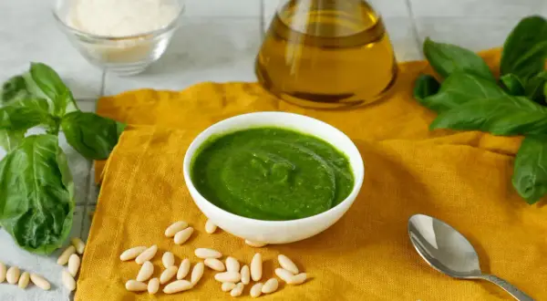 Pesto fresco in una ciotola bianca, con ingredienti per una ricetta pesto semplice: basilico, pinoli, olio d'oliva e parmigiano grattugiato.