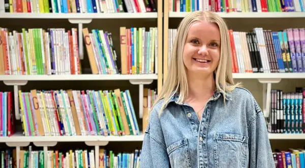 Evelina Persson ler glatt framför bokhyllor fyllda med färgglada böcker.