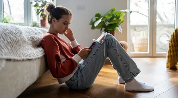 En flicka sitter på golvet, försjunken i sin telefon. Hennes hållning och ansiktsuttryck kan antyda depression hos barn.