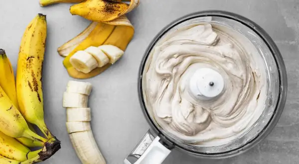 En matberedare fylld med krämig bananfrukt-glass bredvid skalade och skivade bananer.