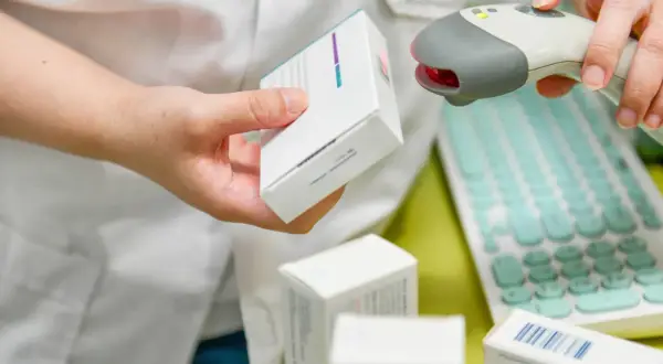 Farmaceuta skanuje lek, pokazując, ile ważna jest recepta. W tle klawiatura i inne opakowania.