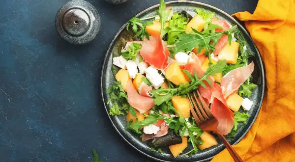Insalata con melone, prosciutto crudo e formaggio fresco, guarnita con rucola. Un piatto estivo perfetto.