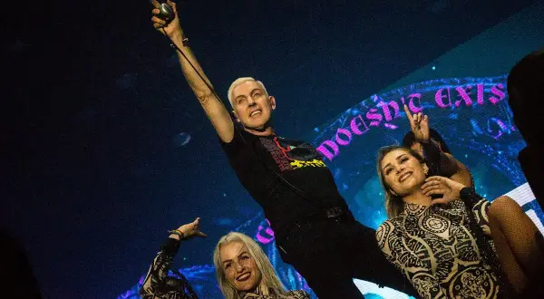 Scooter muzyk, H.P. Baxxter, na scenie z tancerkami, w tle neonowy napis.