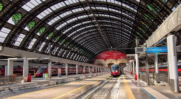 Peron na dworcu w Mediolanie, z pociągami i charakterystycznym, łukowatym zadaszeniem. Idealne miejsce na podróż do Gardy.