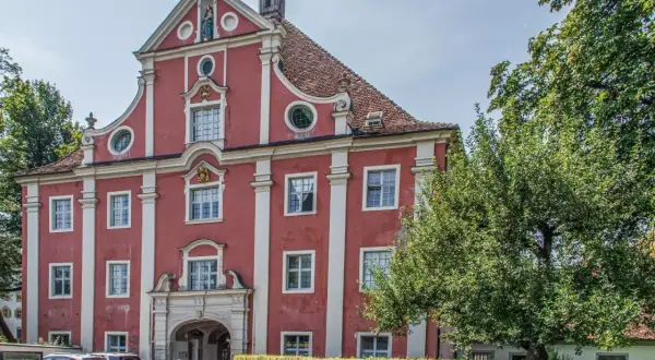 Prachtvolles Gebäude des Klosters Salem mit roter Fassade, weißen Säulen und einem geschwungenen Giebel. Ein Baum spendet Schatten.