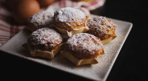 Cinque dolci sardi, spolverati di zucchero a velo, su un piatto bianco. Sullo sfondo, uova e un tessuto a quadri rossi.