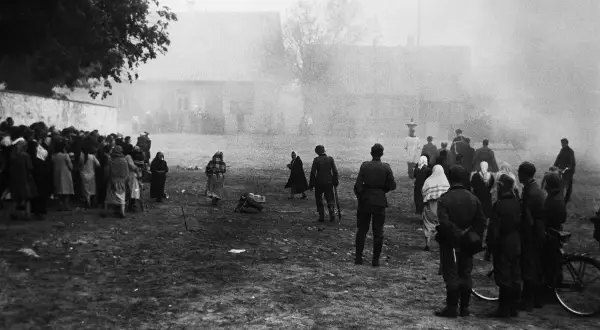 Czarno-białe zdjęcie przedstawia grupę ludzi, w tym żołnierzy, zgromadzonych na otwartym terenie przed budynkami.