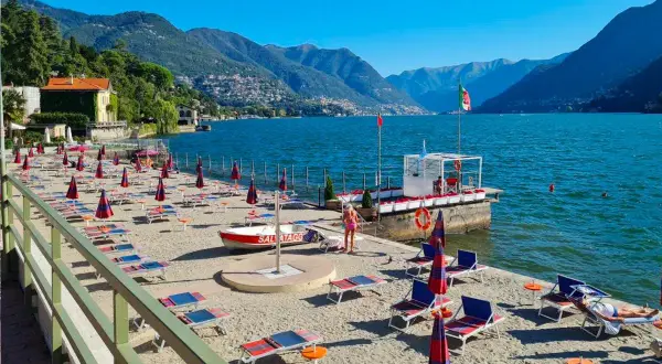 Plaże nad jeziorem Como, z rzędami leżaków i parasoli, czekają na turystów. W tle góry i błękitne niebo.
