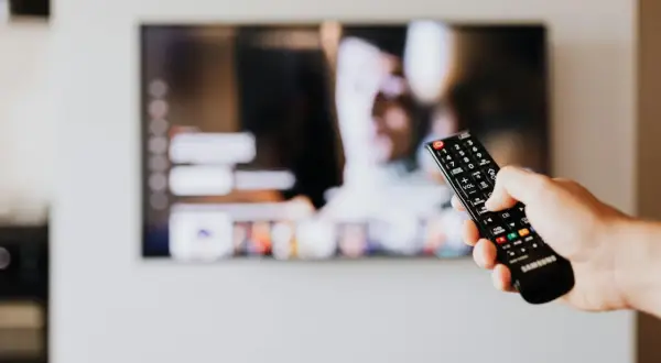 Dłoń trzyma pilot Samsunga, celując w ekran telewizora. Chcesz wiedzieć, jak wyłączyć audiodeskrypcję Samsung?