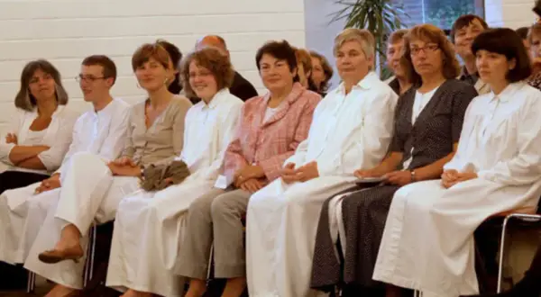 Frauen in weißen Gewändern sitzen nebeneinander. Die Kleidung könnte auf eine religiöse Gemeinschaft hindeuten, wo Baptisten Regeln für Frauen haben.