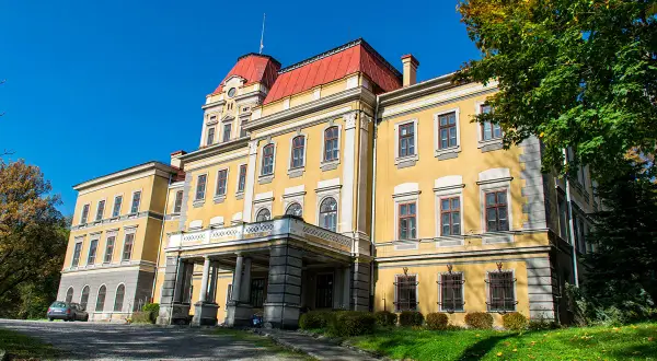 Żółty pałac z czerwonym dachem, otoczony zielenią i jesiennymi drzewami. W oddali widać kończyce wielkie.