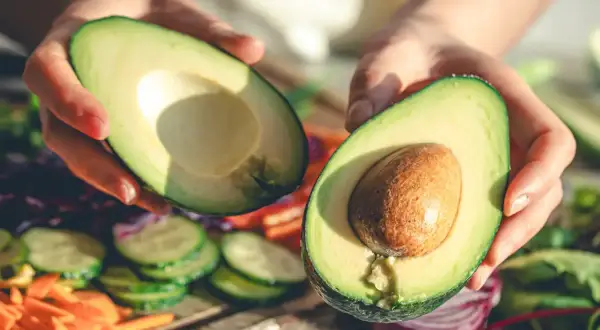 Manos sostienen un aguacate cortado por la mitad, listo para disfrutar. Es la temporada del aguacate en España, con verduras frescas de fondo.