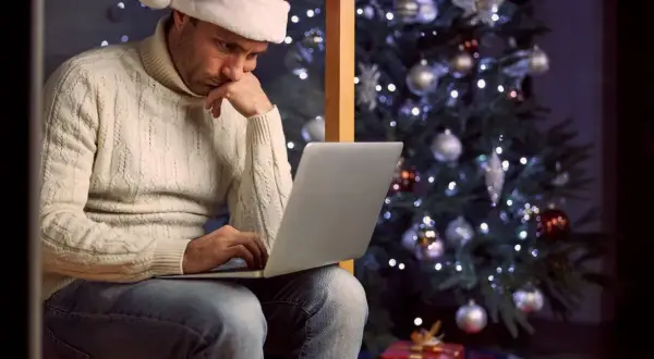 Mężczyzna w czapce Mikołaja siedzi przy choince, pracując na laptopie. Widać prezenty pod choinką. To smutne **święta**.