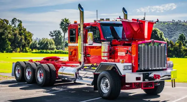 Un impressionnant Kenworth, un camion américain rouge vif, brille sous le soleil. Ses chromes étincelants et ses échappements imposants annoncent sa puissance.