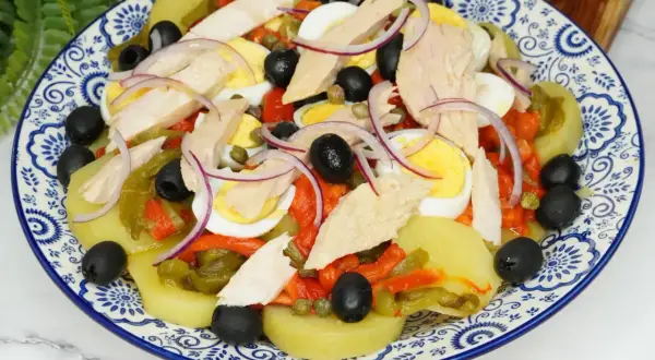Deliciosa ensalada de patata con atún, huevo duro, pimientos asados, aceitunas negras y cebolla morada.