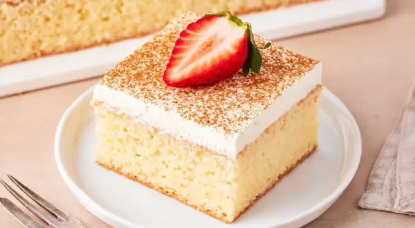 Ein Stück saftiger Tres Leches Kuchen, garniert mit einer Erdbeerhälfte und Zimt, auf einem Teller mit einer Gabel daneben.