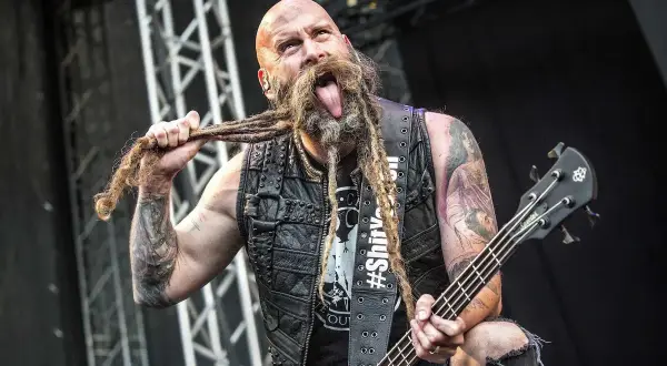 Chris Kael, basista z długą, zaplecioną brodą, wyciąga język podczas koncertu, trzymając gitarę basową.