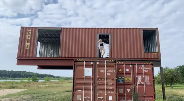 Dom z kontenerów 100m2 cena. Dwa kontenery ustawione jeden na drugim, na górnym stoi kobieta.