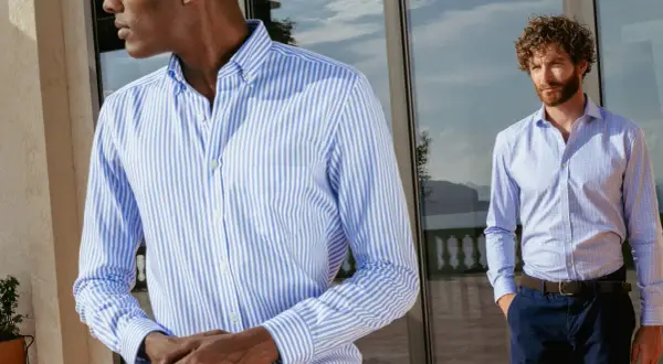 Uomo con camicia personalizzata a righe azzurre e bianche, in piedi vicino a una vetrata.
