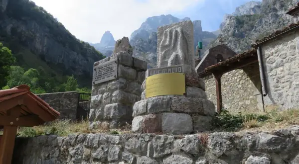 Monumento en piedra con inscripciones y un relieve, con montañas al fondo. El cartel dice "EL CAIN DE ARRIBA".