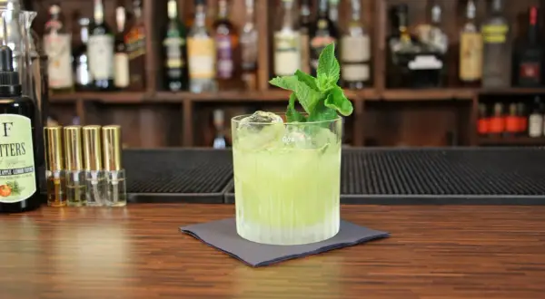 Ein grüner Tequila-Cocktail mit Minze und Eis auf einem Holztresen, im Hintergrund Regale mit Flaschen.