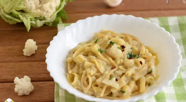 Un piatto di pasta e cavolfiore cremoso, guarnito con prezzemolo e peperoncino. Sullo sfondo, un cavolfiore fresco e uno spicchio d'aglio.