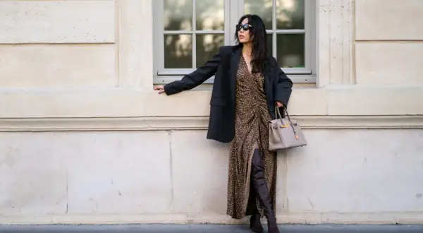 Elegancka kobieta w sukience w panterkę i czarnym blazerze, z beżową torebką i brązowymi kozakami dla niskiej osoby, które optycznie wydłużają nogi.