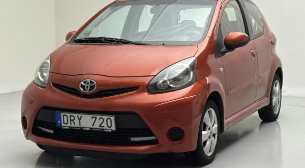 En orange Toyota Aygo med registreringsskylten DRY 720. Bilen verkar ha ett problem med fronten, kanske en buckla eller skada.