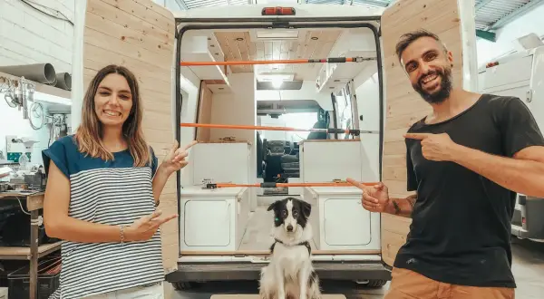 Pareja sonriente y su perro posan junto a una furgoneta camper en proceso de construcción, ¡las mejores furgonetas camper calidad precio!